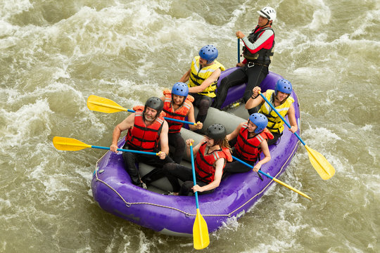 A family of four smiling on a white water raft, navigating through choppy waters with splashes of white foam surrounding them.