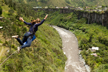 A woman freefalls in mid-air while bungee jumping from a bridge above a winding river and lush green valley in Banos, Ecuador, arms outstretched, capturing the adrenaline of extreme sport. © Ammit