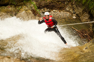 A daring climber rappels down a rugged canyon wall, descending towards a rushing stream and majestic waterfall below.