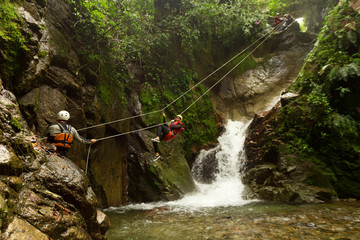 A group of adventurous tourists in Ecuador's Banos Canyon, ziplining over a dangerous waterfall for...