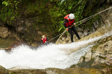 A daring climber rappels down a rocky canyon wall next to a cascading waterfall in the wild wilderness of Ecuador, surrounded by rushing water.