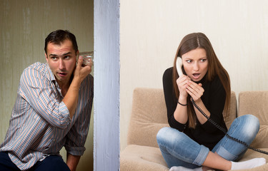 Man with a glass listening to the girl's phone conversation through the wall