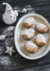 cookies madeleines with powdered sugar on the oval plate, ceramic Santa Claus and Christmas decorations on dark wooden surface