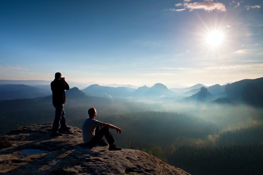 Hiker and photo enthusiast stay with tripod on cliff and thinking. Dreamy fogy landscape, blue misty sunrise in a beautiful valley below