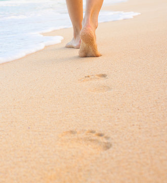 Beach travel - woman walking on sand beach leaving footprints in the sand. Closeup detail of female feet and golden sand.