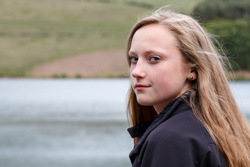 Blonde female teenager looking back over shoulder in front of a lake.  Long hair and slight smile and moody expression.  Bokeh