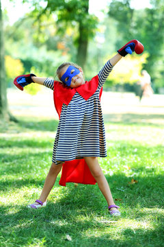 Happy little girl dressed as superhero in boxing gloves at the park