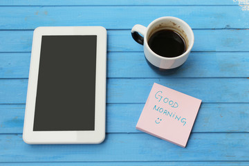 Touch screen tablet computer, cup of coffee, sticky note with good morning message and blue pen - on blue background