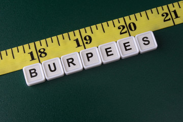 Arrangement of alphabet blocks of word BURPEES with yellow tape measure on the rough dark green background.