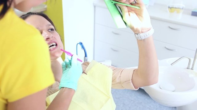 Female dentist showing to her sitting and looking at a mirror female patient how to use dental interproximal brush correctly / camera moving right / close-up