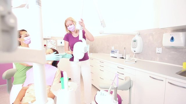 Female dentist assistant giving female dentist x-ray scanner for brown haired female patient sitting in a dental chair / camera moving left
