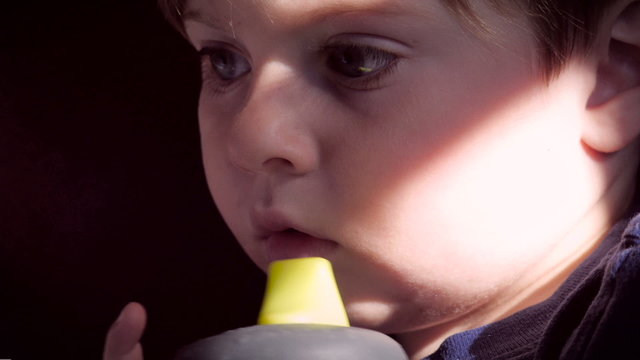 Slow motion of a small child looking concerned with a slash of natural sunlight across his face while holding a sippy cup in his hand.