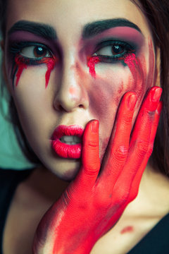 portrait of freak monster with mess dirty colored makeup on her face. crying woman with red bloody tears and hand. halloween concept on green background. studio shot, dark brown eyes.