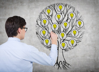 Rear view of young professional who is drawing a tree with light bulbs on the concrete wall. Light...