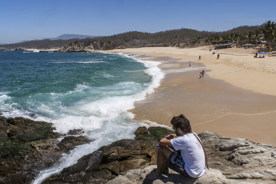 hombre joven pensando y mirando al mar sobre un risco con la playa de fondo