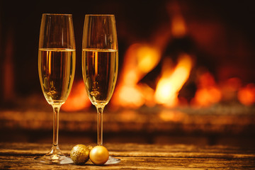 Two glasses of sparkling champagne in front of warm fireplace. Cozy relaxed magical atmosphere in a chalet. Holiday concept. Beautiful background with shimmering wine, decorated with golden baubles.