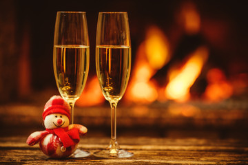 Two glasses of sparkling champagne in front of warm fireplace. Cozy relaxed magical atmosphere in a chalet. Holiday concept. Beautiful background with shimmering wine, decorated with snowman toy.
