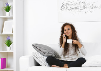 Young woman sitting on comfortable sofa with cup on coffee in hands and speaking by cellphone
