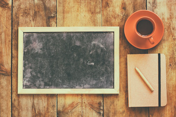 top view image of empty blackboard next to cup of coffee and notebook, over wooden table. image with retro style filter
