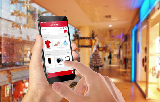 Smart phone online shopping in man hand during Christmas. Shopping center in background. Buy clothes shoes accessories with e commerce web site