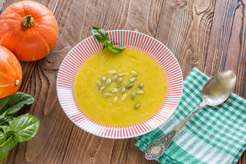 cream soup from pumpkin and other vegetables decorated by basil leaf and pumpkin seeds, basil, pumpkins fruits, salt, pumpkins  seeds  on wooden table with silver spoon on green squared  napkin