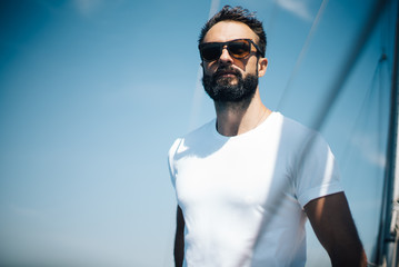 Bearded man wearing sunglasses, standing on a yacht and looking at the horizon