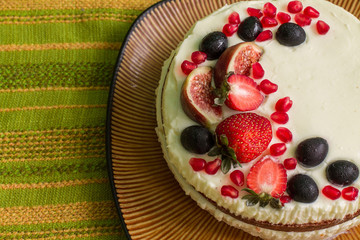 Nude cake with strawberries, figs, grapes and pomegranates on big brown plate. Plate is on green tablecloth with stripes. 