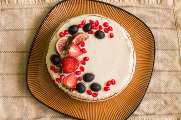 Nude cake with strawberries, figs, grapes and pomegranates on big brown plate. Plate is on green tablecloth with stripes. 