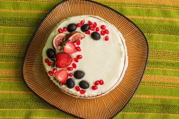 Naked cake with strawberries, figs, grapes and pomegranates on big brown plate. Plate is on green tablecloth with stripes. Aerial view.