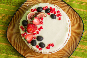 Naked cake with strawberries, figs, grapes and pomegranates on big brown plate. Plate is on green tablecloth with stripes. Aerial view.