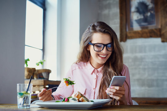 Young woman eating salad at restaurant and texting on smartphone