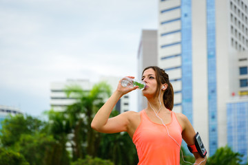 Fitness woman drinking water from bottle during running or workout rest at city park. Female athlete taking a rest for hydration.