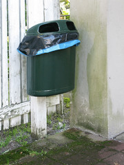 Ugly plastic waste paper basket in mossy green corner outside a residential building in need of renovation.