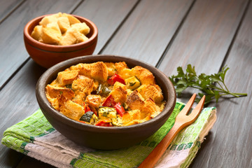 Savory baked vegetarian bread pudding made of zucchini, bell pepper, tomato, thyme, parsley, photographed with natural light (Selective Focus, Focus on the zucchini pieces in the front)