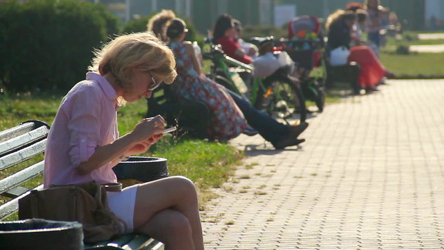 Beautiful young woman using smart phone on bench in park, tapping touch screen