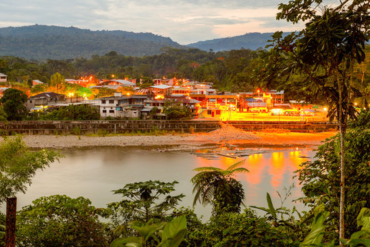 A serene riverside view of the Misahualli port at twilight, warm golden lights reflecting on the calm Napo river surrounded by lush greenery and distant hills in the Amazon basin of Ecuador.