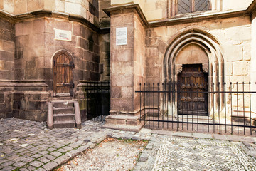 Fototapeta premium A close-up of the ancient Gothic stone facade of the Black Church in Brasov, Romania, a weathered wooden door in a pointed arch behind an iron fence and cobblestone courtyard.