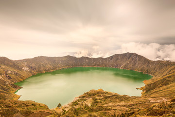 Naklejka premium The serene emerald-green Quilotoa crater lake in a volcanic caldera surrounded by arid rocky mountains under an overcast sky in the Andes of Ecuador, a scene of natural wonder.