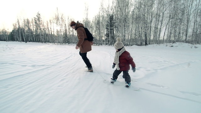 Little boy learning to ride a snowskate pulled by his young father 
