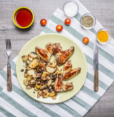 Fried delicious chicken wings with fried potatoes spicy chili sauce and cherry tomatoes and seasoned with a fork and knife on a striped napkin on rustic wooden background  top view close up