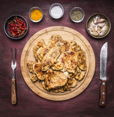 lot of fried appetizing chicken steaks with herbs hot pepper and garlic with a fork and knife on a round cutting board on rustic wooden background top view