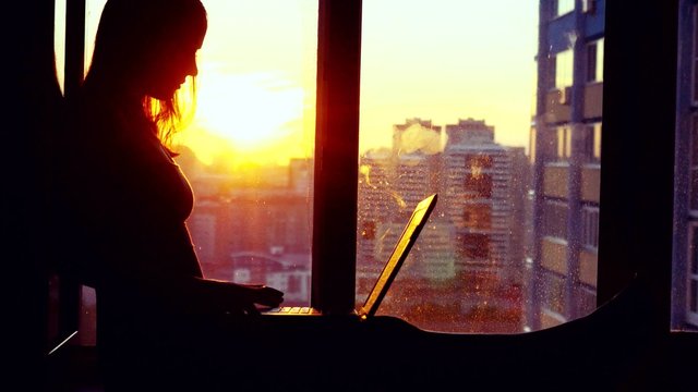 Beautiful young brunette business woman working on her laptop during sunset. Girl sitting on the window at modern building blurred background in city