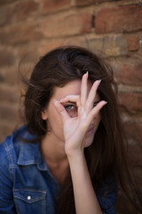 portrait of young, youth, pretty,funny, pale, happy ,smileing girl with long brown hair and a birthmark on face beside brick wall wearing jeans blue shirt looking in camera through fingers 