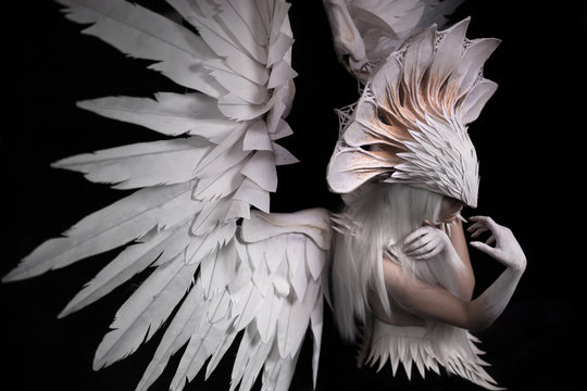  a portrait of a young girl and a white wig , which carries a large white mask and a large white wings. feathers costume and hands painted in white, head down , looking sideways, thoughtful.