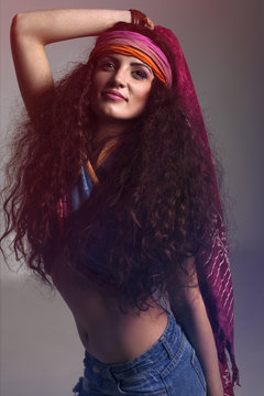 Young hippie girl  with long, brown, curly hair,  wearing colorful scarf around forehead ,her hand is behind head, wearing colorful top over her breasts, seductively smileing 