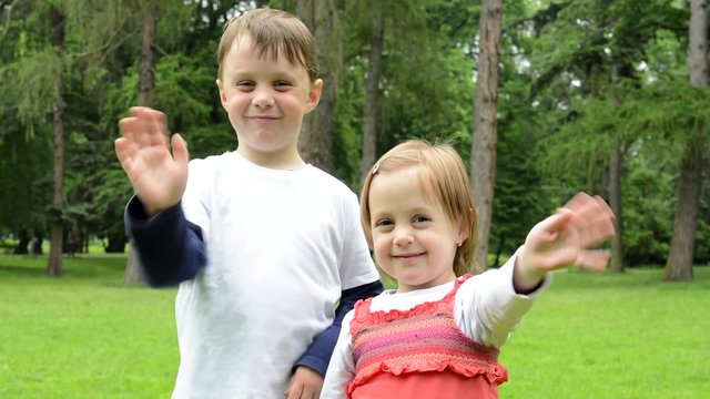 childrens (young boy and little girl (siblings)) wave with hands and smile to camera  - park