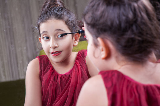 Small beautiful arab middle eastern girl with pretty red dress and lips doing makeup carefully at home in mirror. 8-10 years with dark brwon hairs and dark eyes.