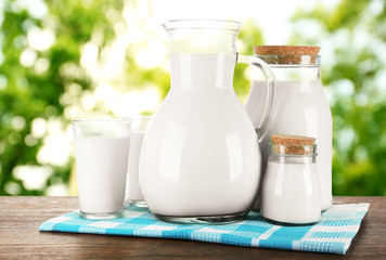 Pitcher, jars and glasses of milk on wooden table, on nature background