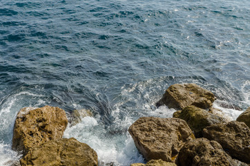 close-up of waves of the mediterranean sea water crushing into reddish rocks of the shore on a sunny day
