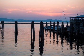 Fototapeta premium pink skies of sunset light up the lake and boathouse with pier, sailboat, and pylons in foreground with the Adirondack mountains across the lake 